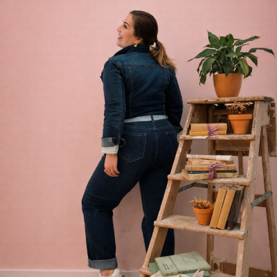 Mulher com roupa de ganga azul escura e sapatilhas brancas junto a escada de madeira com livros e plantas