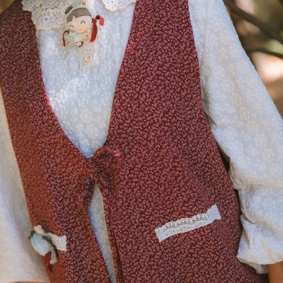 Colete vermelho floral com renda e bonecos de tecido sobre camisa branca com padrão floral e gola rendada