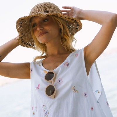 Mulher com vestido branco floral, óculos de sol bege, chapéu de palha castanho claro ao ar livre de frente para o mar.