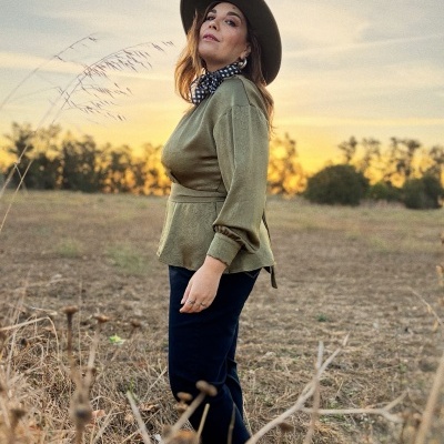 Mulher jovem com camisa verde, calças escuras, lenço preto com padrão branco e chapéu castanho em campo ao pôr do sol