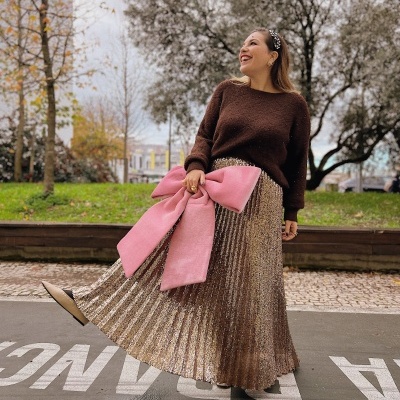 Mulher com camisola castanha, saia dourada plissada e clutch rosa com laço grande no parque.