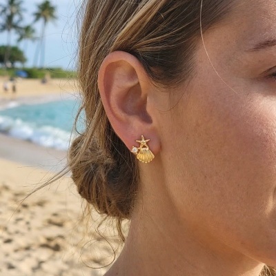 Brinco dourado com design de estrela-do-mar, concha e pedra branca na orelha de mulher na praia