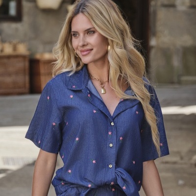Mulher com camisa azul de manga curta com flores rosa e calças com cinto, em rua com pano de fundo de parede de pedra e acessórios pendurados.