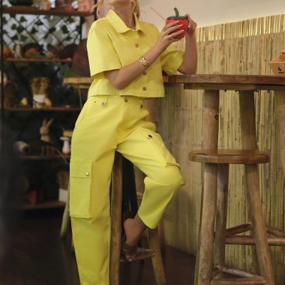 Mulher vestindo conjunto amarelo com calças cargo e camisa, sentada em banco de madeira, segurando caneca vermelha.