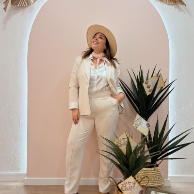 Mulher com fato creme e chapéu bege junto a plantas em vasos de palha junto a parede rosa com arco