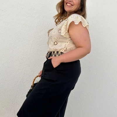 Mulher com blusa branca rendada e saia preta com cinto posando contra parede branca