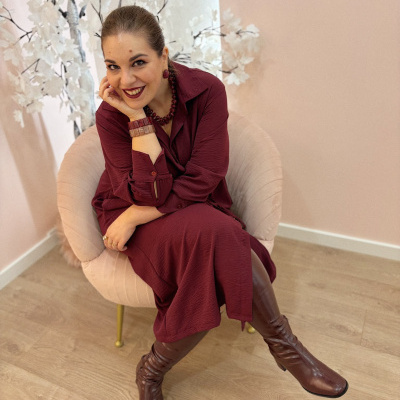 Mulher sentada em poltrona rosa usando vestido vinho e botas castanhas.
