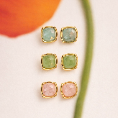 Três pares de brincos dourados com pedras facetadas azul claro, verde e rosa sobre fundo branco e flor laranja