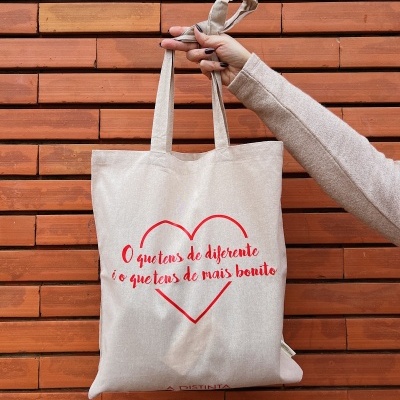 Mala tote bege com texto vermelho e coração contra parede de tijolos vermelhos