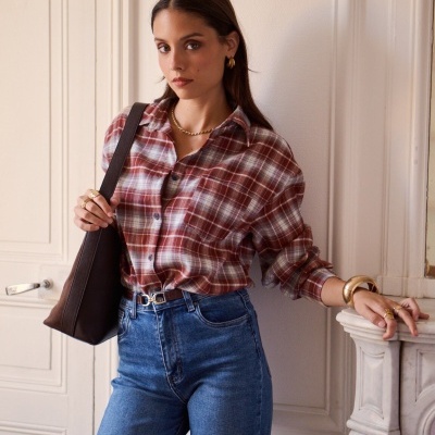 Mulher com camisa xadrez vermelha e branca, jeans azuis e mala castanha ao ombro junto a parede interior