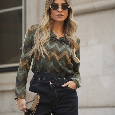 Mulher com camisola padrão zigzag, calças de ganga pretas, óculos de sol e mala bege com correntes douradas