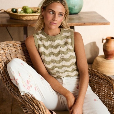 Blusa de malha verde claro e branca com calças brancas bordadas sentada numa cadeira de vime em ambiente rústico.