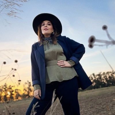 Mulher com jaqueta azul, blusa verde, chapéu preto e lenço ao ar livre ao pôr do sol