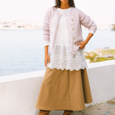 Mulher com camisola branca, cárdigan rosa bordado, saia castanha e botas castanhas perto do mar