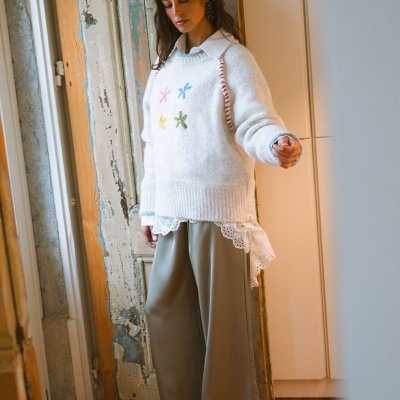 Mulher com suéter branco e flores bordadas, camisa branca com renda e calças largas cinza esverdeado