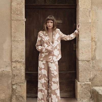 Conjunto feminino bege com flores brancas, blazer e calça larga, modelo junto a porta de madeira e parede de pedra