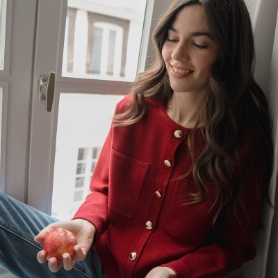 Mulher de cardigan vermelho com botões dourados a segurar uma maçã e sentada numa janela