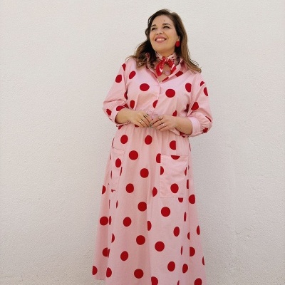Mulher com vestido rosa de bolinhas vermelhas e lenço ao pescoço, junto de parede branca