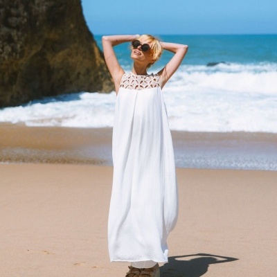 Vestido longo branco com renda floral numa praia com mar e rochas.