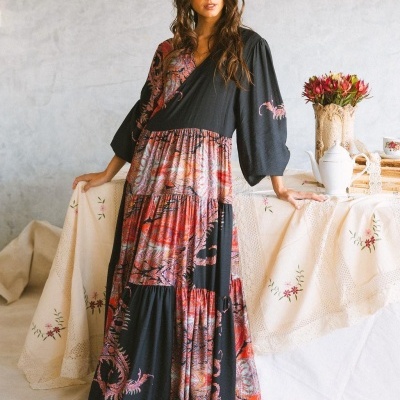 Vestido comprido floral em preto, vermelho e lilás com mangas 3/4 junto a mesa com toalha bordada e flores.