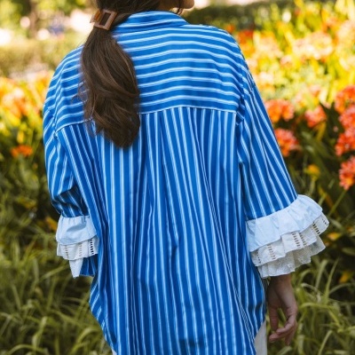 Camisa azul às riscas brancas com folhos nas mangas e cabelo preso com presilha castanha, vista de costas em jardim florido.