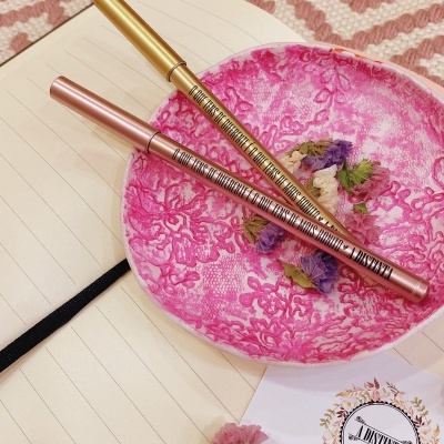 Dois lápis delineadores em um prato rosa decorativo sobre caderno aberto e papel branco com selo e flores secas