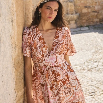 Vestido rosa e castanho com padrão floral, mulher encostada em parede de pedra