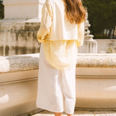 Mulher vista de costas com camisa amarela clara com pequenos corações, colete branco, calças brancas e sapatos castanhos, ao ar livre.