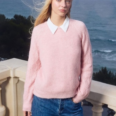 Mulher com camisola rosa de malha e gola branca vestindo jeans azuis, em varanda com vista para o mar
