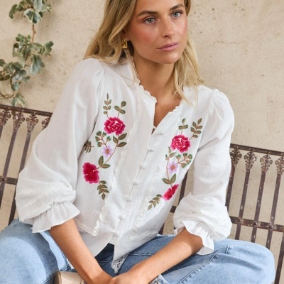 Blusa branca com bordados florais vermelhos e rosa e botões, combinada com jeans azuis claros, modelo sentada num banco de metal.