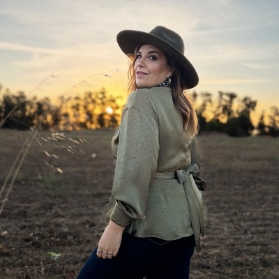 Mulher com casaco verde-oliva e chapéu castanho em campo ao pôr do sol.