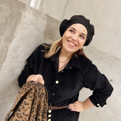 Mulher a sorrir com boina preta e casaco com padrão animal print em fundo de parede de cimento.