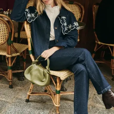 Mulher sentada com roupa de ganga, botas castanhas e mala verde numa cadeira de madeira e palhinha.