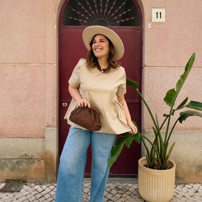 Mulher com roupa casual bege e azul junto a porta bordô e planta em vaso