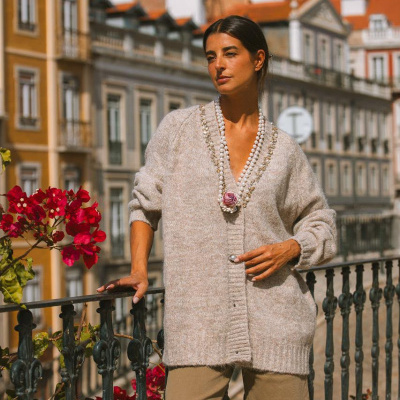 Mulher com cardigã cinza e colar de pérolas em varanda urbana com flores vermelhas