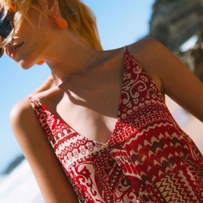 Vestido vermelho e branco com padrão étnico em tecido leve, usado por mulher com brincos alaranjados e óculos de sol sob luz solar junto ao mar
