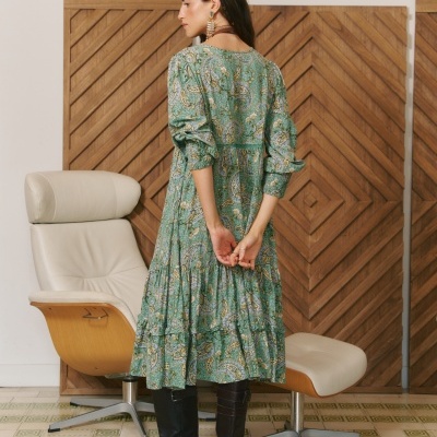 Mulher de costas com vestido longo verde estampado e botas pretas em sala com cadeira e parede de madeira.