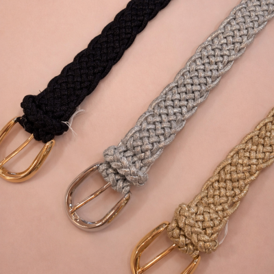 Três cintos trançados em tecidos brilhantes preto, prata e dourado com fivelas metálicas em fundo rosa