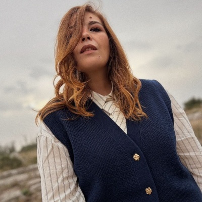 Mulher com colete azul e camisa branca às riscas, céu nublado ao fundo