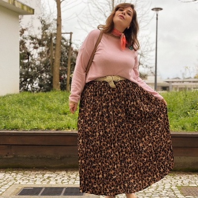 Mulher com saia plissada animal print, camisola rosa, lenço coral, cinto amarelo e sandálias castanhas