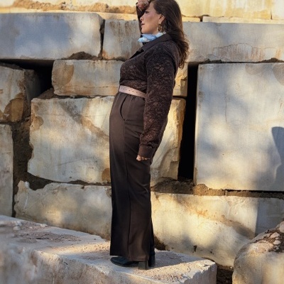 Mulher de perfil com blusa de renda castanha, calças castanhas e brincos grandes ao ar livre, rodeada por pedras grandes claras.