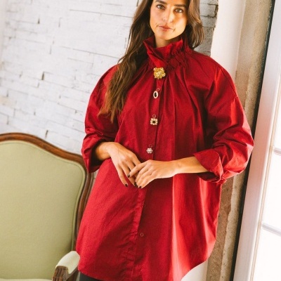 Mulher com blusa vermelha de manga comprida e botões decorativos junto a parede branca e cadeira verde