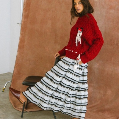 Mulher com camisola vermelha e saia longa branca com riscas pretas, sapatos castanhos em cadeira preta