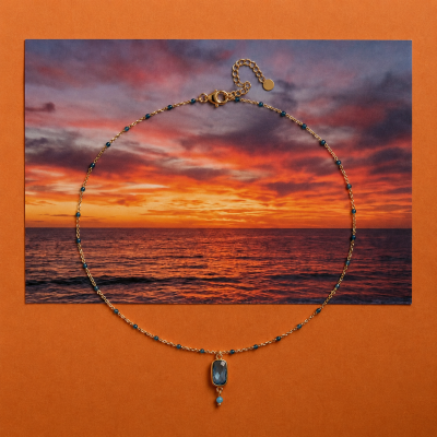 Colar dourado com pendente azul sobre imagem de pôr do sol no mar e fundo laranja.