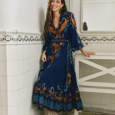 Mulher com vestido azul escuro estampado e botas creme com franjas, em divisão com azulejos brancos e mesa branca