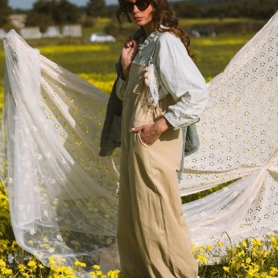 Mulher com macacão castanho claro e camiseta branca em campo com flores amarelas e pano branco decorativo