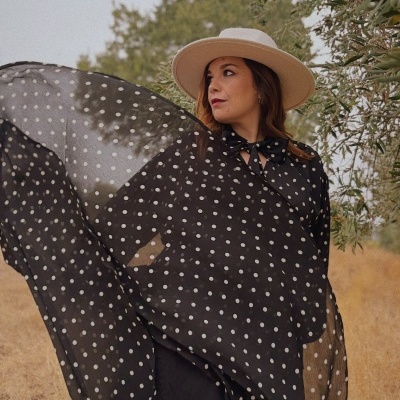 Blusa preta de bolinhas brancas com laço no decote e chapéu bege em ambiente natural.