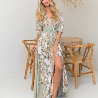 Vestido longo floral verde e rosa com botões e cordão na cintura, usado por mulher junto a mesa de madeira.