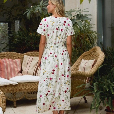 Vestido branco com padrão floral colorido, visto pelas costas, usado por mulher em ambiente com móveis de rattan e plantas.