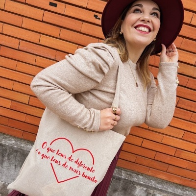 Mulher com sacola de tecido bege com texto vermelho, blusa bege, saia plissada vinho e chapéu vinho, junto a parede de tijolos laranja